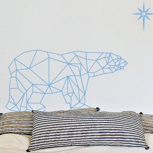 Puede incluir: Un adhesivo de pared de oso polar geométrico azul claro con una estrella encima. La calcomanía está en una pared blanca sobre una cama con ropa de cama blanca y almohadas a rayas.