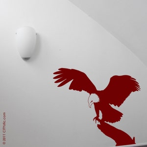 Könnte beinhalten: Eine rote Silhouette eines Adlers mit ausgebreiteten Flügeln, der auf einem Ast landet. Der Adler blickt nach rechts. Der Hintergrund ist eine weiße Wand.