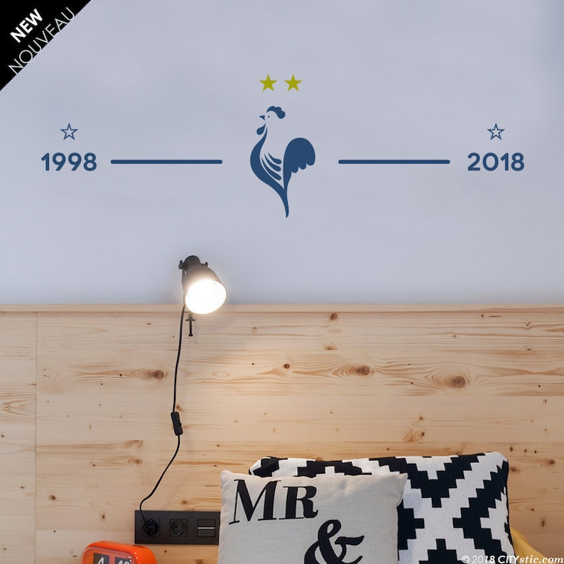 Puede incluir: Una pared blanca con una silueta de gallo azul y dos estrellas doradas encima. El gallo est&aacute; centrado entre los a&ntilde;os 1998 y 2018, que est&aacute;n escritos a ambos lados del gallo. La pared est&aacute; sobre una cabecera de madera con una l&aacute;mpara y almohadas.