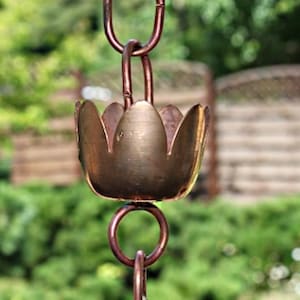 Op de afbeelding: Een koperen regenketting met een decoratieve bloemvormige beker. De ketting is gemaakt van verbonden ringen en hangt aan een haak. De achtergrond is wazig en toont groen gebladerte en een houten hek.