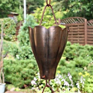 Op de afbeelding: Een hangende koperen regenkoord met een uitlopend, geschulpt bekervormig ontwerp. De ketting is opgehangen aan een koperdraad. De achtergrond is een vage groene omgeving en een houten hek. Decoratief item voor buiten.