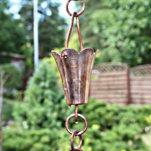 Pode incluir: Uma corrente de chuva de cobre com um design de copo canelado decorativo. A corrente de chuva é suspensa por um anel e possui vários anéis de conexão. O fundo é uma vista desfocada de um jardim com folhagem verde e uma cerca de madeira.