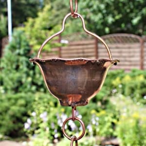 Op de afbeelding: Een koperen regenkoord met een decoratieve komvormige beker en een ophangring. De regenkoord hangt voor een vage groene achtergrond van bomen en een houten hek. De kom heeft een geschulpte rand.