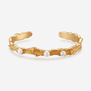 May include: Gold-toned cuff bracelet with a textured, organic design. The bracelet features three small, round, white pearls set along the front. The open design allows for easy wear and removal. A stylish piece of jewelry.
