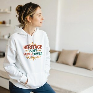 May include: A white hoodie featuring the text "HERITAGE IS MY SUPERPOWER" in red, green, and gold. The hoodie has a front pocket and a drawstring hood. The person in the image is wearing the hoodie.