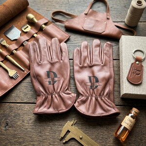 May include: A pair of tan leather gloves featuring a black monogram with the letter D and the text Dillo. The set includes a leather tool roll with brass tools, a leather apron, a leather keychain, a spool of thread, and a bottle of Dillo Care Oil.