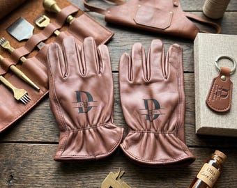 May include: A pair of tan leather gloves featuring a black monogram with the letter D and the text Dillo. The set includes a leather tool roll with brass tools, a leather apron, a leather keychain, a spool of thread, and a bottle of Dillo Care Oil.