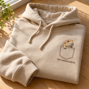 May include: A beige hooded sweatshirt featuring an embroidered graphic of a bearded dragon peeking out of a chest pocket. Below the pocket, the text reads "TINY DRAGON KEEPER" in dark brown lettering. The hoodie includes a drawstring hood.