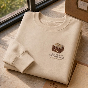 May include: A beige crewneck sweatshirt featuring an embroidered graphic of a brown file box containing documents and a key. Below the box, the text reads: GUARDIAN OF THE PAPER TRAIL RECORDS MANAGER. The sweatshirt has long sleeves with ribbed cuffs.