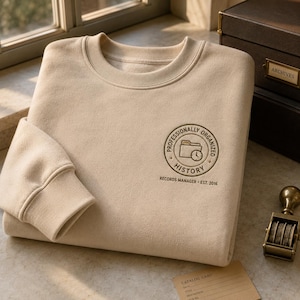 May include: A beige crewneck sweatshirt featuring a circular embroidered emblem on the chest. The emblem displays a file folder and clock icon with the text "PROFESSIONALLY ORGANIZED HISTORY" and "RECORDS MANAGER - EST. 2016" printed below it.
