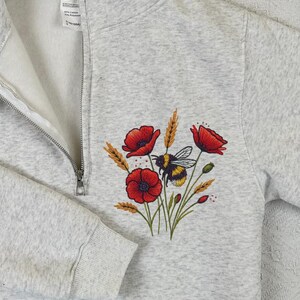 Peut inclure: Un sweat-shirt gris clair à fermeture éclair quart de zip avec un motif brodé de coquelicots rouges, de blé et d'un bourdon. Le sweat-shirt a un intérieur blanc et une fermeture éclair argentée.