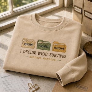 May include: A beige crewneck sweatshirt featuring embroidered file folder icons labeled RETAIN, ARCHIVE, and DESTROY. Below the icons, the text reads I DECIDE WHAT SURVIVES and RECORDS MANAGER in dark brown and green thread, centered on the chest.