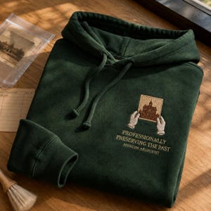 May include: A dark forest green hooded sweatshirt featuring an embroidered graphic on the chest. The design depicts two hands holding a framed building illustration above the text "PROFESSIONALLY PRESERVING THE PAST MUSEUM ARCHIVIST."