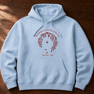 May include: A light blue hooded sweatshirt featuring a dark red graphic on the chest. The design includes a circular branch wreath, a four-pointed star, and the text "BE CAREFUL WHAT YOU WISH FOR" above and "ONE WISH • 2026" below the graphic.