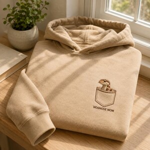 Peut inclure: Un sweat-shirt à capuche beige avec un motif brodé sur la poitrine. Le dessin représente un petit serpent hognose brun sortant d'un contour de poche cousu, avec le texte HOGNOSE MOM brodé en lettres brunes juste en dessous.