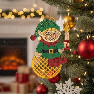 Może przedstawiać: Haftowana ozdoba świąteczna przedstawiająca elfa trzymającego świecę. Elf ma zielony kapelusz i kurtkę z czerwonymi akcentami. Ozdoba wisi na choince z czerwonymi i złotymi bombkami. Widoczny tekst "ORNAMENT EMBROIDERY" i "3" TO 6" SIZES".