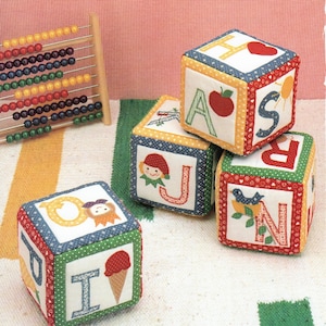 Puede incluir: Cuatro bloques de tela coloridos con las letras A, H, J y N, cada uno con un diseño diferente. Los bloques están apilados sobre una alfombra rayada blanca y verde.