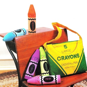 Może przedstawiać: Żółta torba płócienna z zielonym wykończeniem i napisem "5 Dużych Kredki Każda 10 in x 2 1/2 in Nietoksyczna" z przodu. Pięć filcowych kredek w niebieskim, pomarańczowym, zielonym, fioletowym i różowym schemacie kolorów leży na drewnianym biurku.