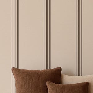 May include: A neutral-toned living room scene featuring a white sofa with several throw pillows in shades of brown and beige. A dark brown coffee table holds two decorative bowls and a small vase. The background has a striped wallpaper.
