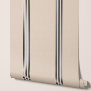 May include: A roll of wallpaper featuring a cream-colored background with three vertical navy blue stripes. The wallpaper is partially unrolled, revealing the pattern and texture of the material. The design is simple and classic.