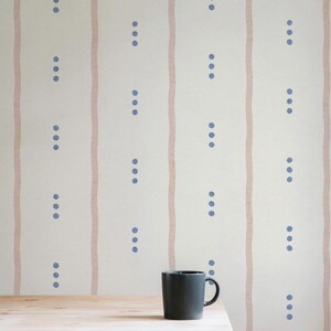 May include: A close-up of a wallpaper design featuring vertical, light pink stripes and rows of blue dots. A dark gray mug sits on a wooden surface in the foreground. The wallpaper has a simple, minimalist aesthetic.