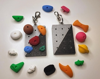 May include: Two rectangular keychains with climbing hold designs. Each keychain has a silver and black diagonal pattern and is adorned with colorful, miniature climbing holds in various shapes and sizes. The holds are in shades of green, orange, red, blue, and white.