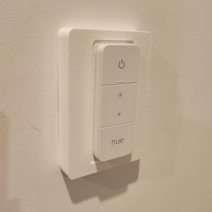 May include: A white smart light switch with three buttons and the brand name "hue" printed on the bottom. The top button has a power symbol, and the middle two buttons have light-related symbols. The switch is mounted on a white wall.