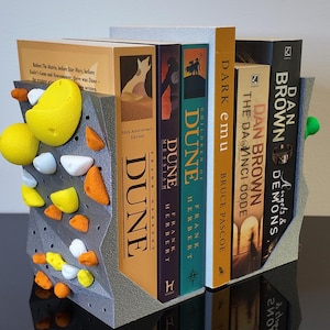 May include: A gray bookend shaped like a rock climbing wall with textured surfaces and small, colorful climbing holds in yellow, orange, and white. The bookend supports a row of books including titles by Frank Herbert and Dan Brown.