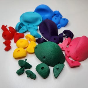 May include: A selection of multi-coloured climbing holds in diverse shapes and sizes. The holds are in shades of red, yellow, green, blue, purple, and pink. Designed for indoor or outdoor rock climbing and bouldering.