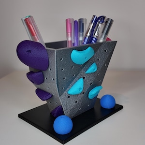 Puede incluir: Un portalápices gris con presas de escalada moradas y turquesas. El soporte está lleno de bolígrafos y se asienta sobre una base negra. Dos esferas azules están en la base del soporte.