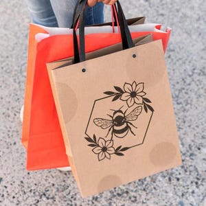 May include: A person holding three shopping bags. The front bag is tan with a black bee and floral design inside a hexagon. The other bags are orange and brown with black handles.