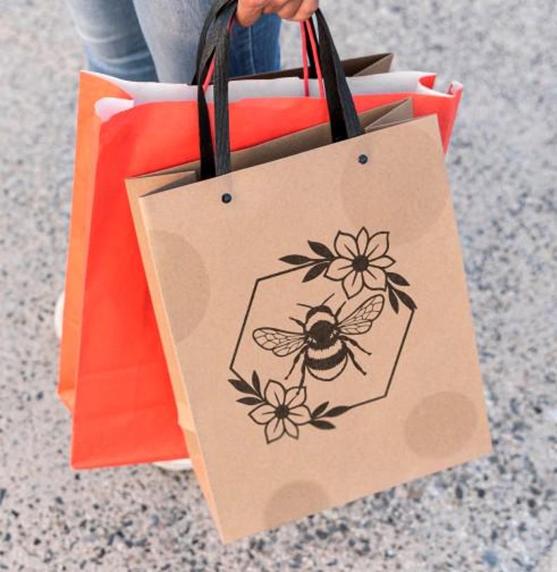 May include: A person holding three shopping bags. The front bag is tan with a black bee and floral design inside a hexagon. The other bags are orange and brown with black handles.