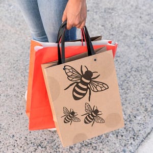 May include: A brown paper shopping bag with black handles featuring a large bee illustration at the top and two smaller bee illustrations below it, layered in front of a bright orange shopping bag with black handles.