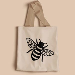 May include: A light beige tote bag featuring a black graphic illustration of a honeybee centered on the front. The bag has two matching light beige fabric handles attached to the top, providing a simple and functional design for carrying items.