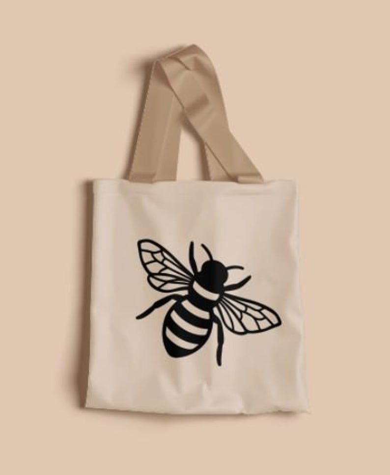 May include: A light beige tote bag featuring a black graphic illustration of a honeybee centered on the front. The bag has two matching light beige fabric handles attached to the top, providing a simple and functional design for carrying items.
