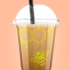 May include: A clear plastic cup filled with a light brown beverage, topped with a clear lid and a black straw. The cup features a yellow honeycomb pattern and a yellow bee graphic. The background is a solid peach color.