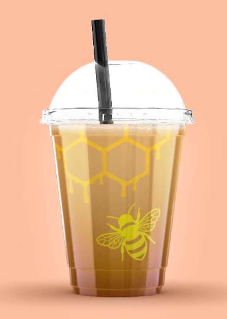 May include: A clear plastic cup filled with a light brown beverage, topped with a clear lid and a black straw. The cup features a yellow honeycomb pattern and a yellow bee graphic. The background is a solid peach color.