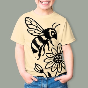 May include: A light yellow short-sleeve t-shirt featuring a large black graphic print of a bee in flight above a daisy flower. The design is centered on the front of the garment, showcasing a bold, high-contrast illustration of the insect.