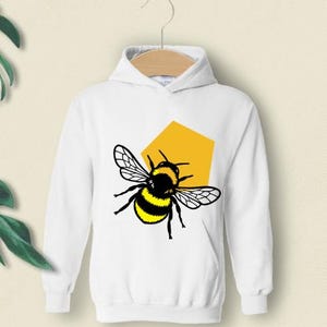 May include: A white long-sleeve hooded sweatshirt featuring a graphic print of a bumblebee with black and yellow stripes and translucent wings, positioned in front of a solid yellow pentagonal shape on the center of the chest.