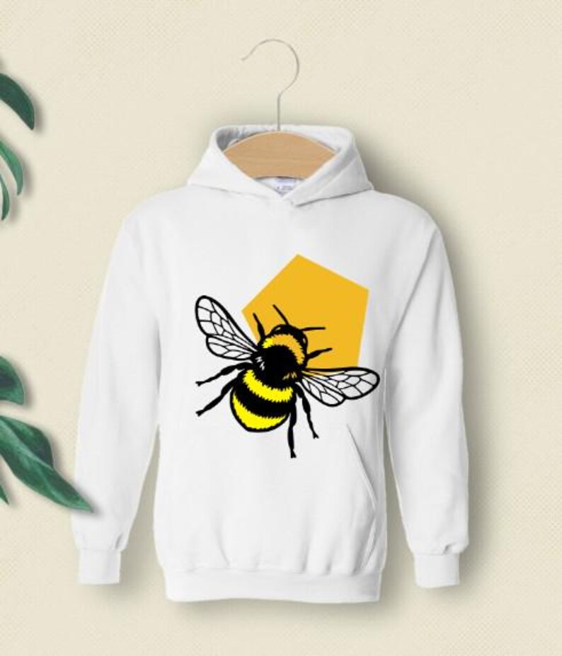 May include: A white long-sleeve hooded sweatshirt featuring a graphic print of a bumblebee with black and yellow stripes and translucent wings, positioned in front of a solid yellow pentagonal shape on the center of the chest.