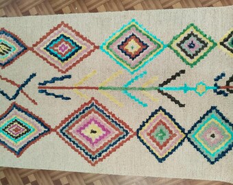May include: Beige woven rug with a geometric pattern. The design features rows of colorful diamond shapes in shades of pink, blue, green, and yellow. An arrow-like motif runs through the center of the rug.