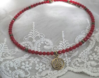 May include: A red beaded choker necklace with a gold-colored pendant featuring a trident design. The necklace is made of small, round red beads and has gold-colored accents. The pendant hangs from the center of the necklace.