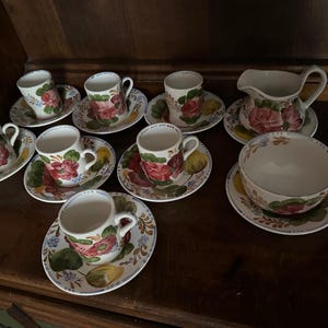 Puede incluir: Un juego de tazas de té y platillos vintage con flores, con una base crema y diseños florales coloridos. El juego incluye una jarra de crema con un diseño a juego.