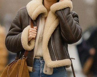 Puede incluir: Una chaqueta de cuero marrón oscuro con un grueso forro de borreguillo color crema, cuello ancho y cremalleras plateadas. La chaqueta se combina con unos vaqueros azul claro y un bolso tipo cubo de ante marrón suave con correa.