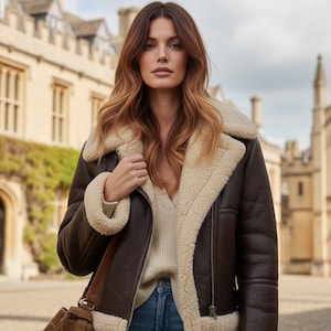 Puede incluir: Chaqueta de cuero marrón con forro y ribete de borrego color crema. La chaqueta tiene un diseño de cremallera asimétrica y un estilo clásico. La persona lleva un jersey color crema y vaqueros azules.