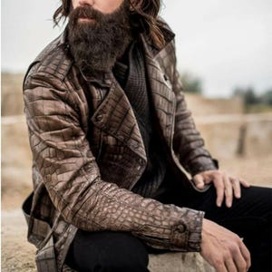 Puede incluir: Chaqueta de cuero marrón con textura de piel de cocodrilo. La chaqueta tiene cuello con solapas, puños abotonados y diseño cruzado. Se lleva sobre un jersey gris oscuro y pantalones negros.