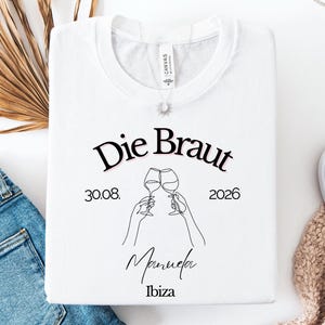 May include: A white t-shirt featuring a black line art graphic of two hands clinking wine glasses. The text reads Die Braut, 30.08. 2026, Manuela, and Ibiza. The design is centered on the chest, with a small brand tag visible at the inner neckline.