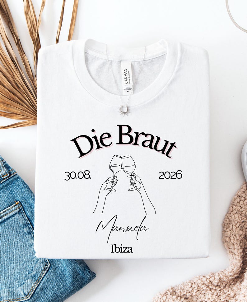 May include: A white t-shirt featuring a black line art graphic of two hands clinking wine glasses. The text reads Die Braut, 30.08. 2026, Manuela, and Ibiza. The design is centered on the chest, with a small brand tag visible at the inner neckline.