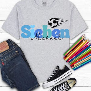 May include: A light gray t-shirt with the name "Sieben Michael" in blue and black, a soccer ball graphic, dark blue jeans, black and white sneakers, and a set of colored pencils.