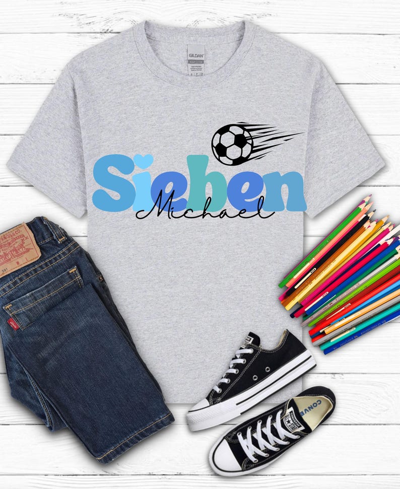 May include: A light gray t-shirt with the name "Sieben Michael" in blue and black, a soccer ball graphic, dark blue jeans, black and white sneakers, and a set of colored pencils.